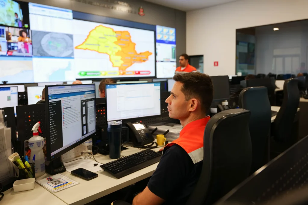 Agente da Defesa Civil diante de telões com mapa de alerta do estado de São Paulo