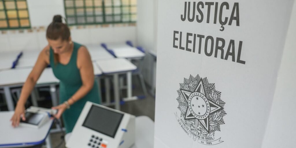 Mesária prepara urna eletrônica em sala de aula ao lado de cabine da Justiça Eleitoral