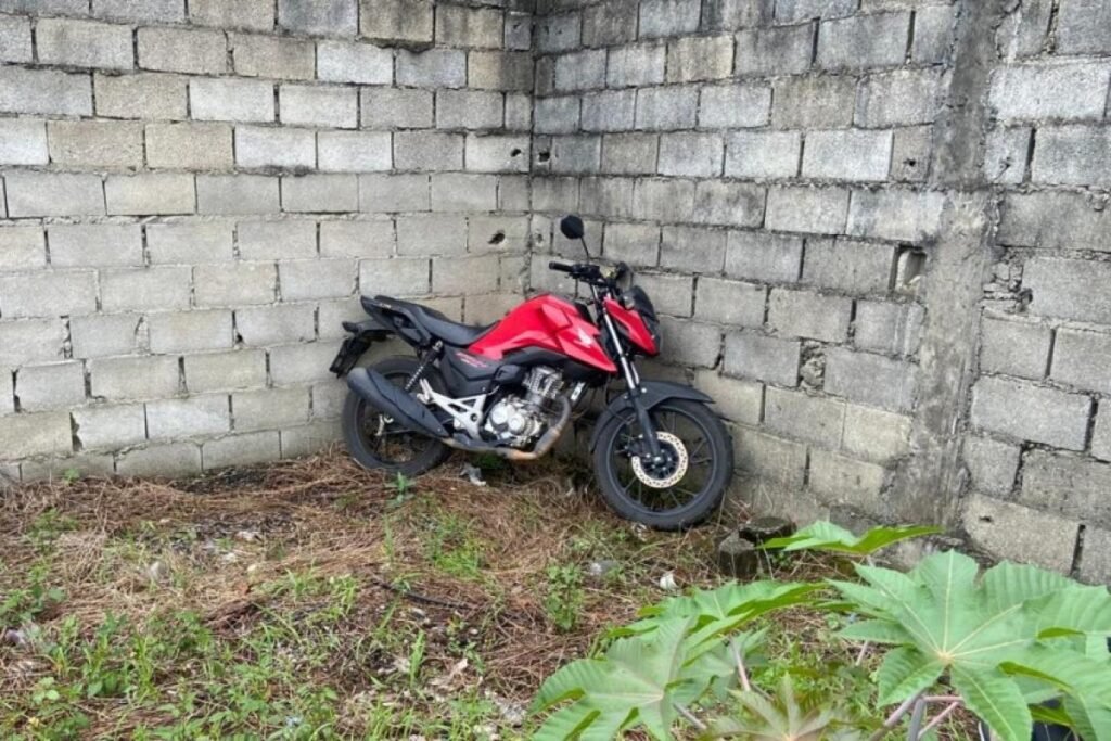 Motocicleta vermelha encostada em muro de blocos, em terreno com mato